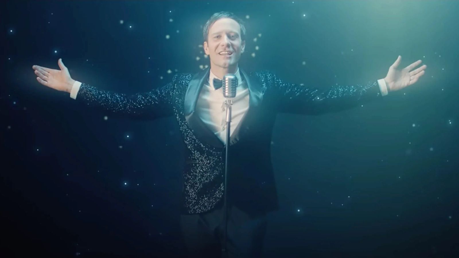 Klemen Slakonja in a glittering tuxedo, arms spread wide at a vintage standing microphone, bathed in deep teal theatrical light against a starfield backdrop