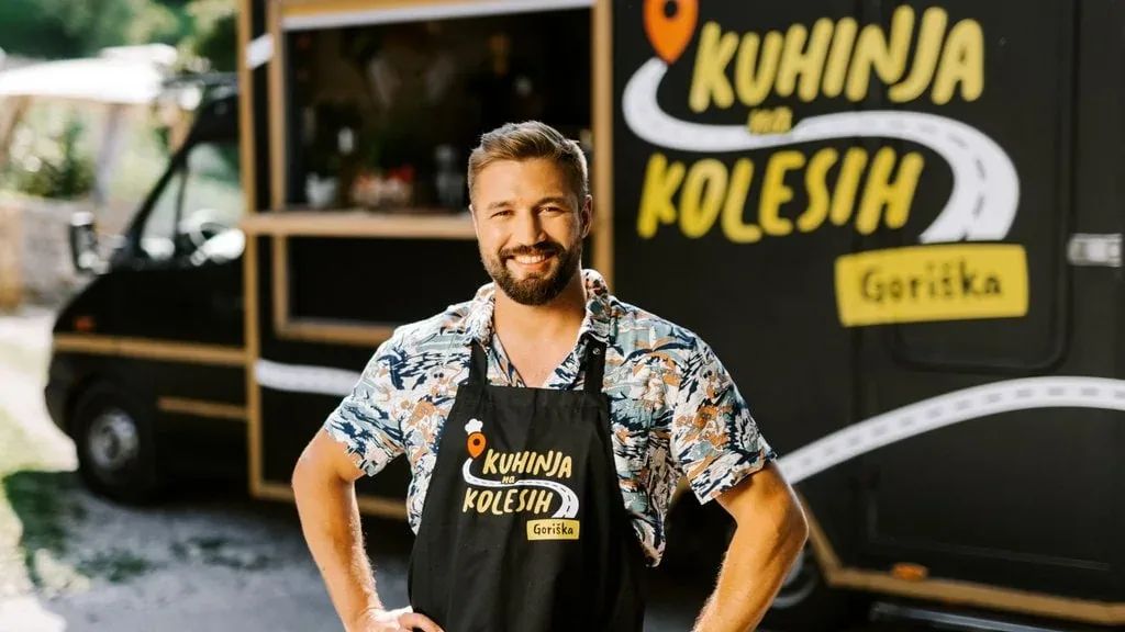 Filip Flisar standing in front of the black Kuhinja na kolesih mobile-kitchen van, wearing the show's apron branded for the Goriška season