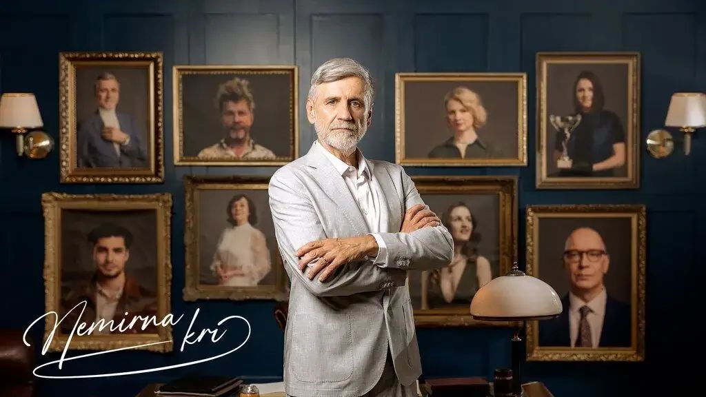 Lead actor of Nemirna kri stands arms crossed before a gallery wall of framed character portraits in an elegant wood-panelled room, the show title in cursive at lower left