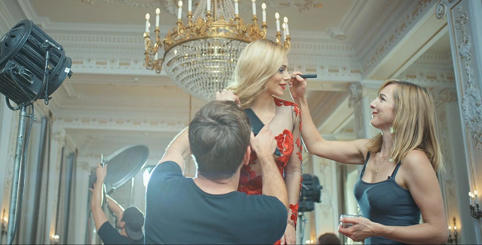 Behind-the-scenes on a commercial shoot — a makeup artist and stylist attend to Katarina Cas in a red floral gown beneath an ornate gilded chandelier in a grand ballroom