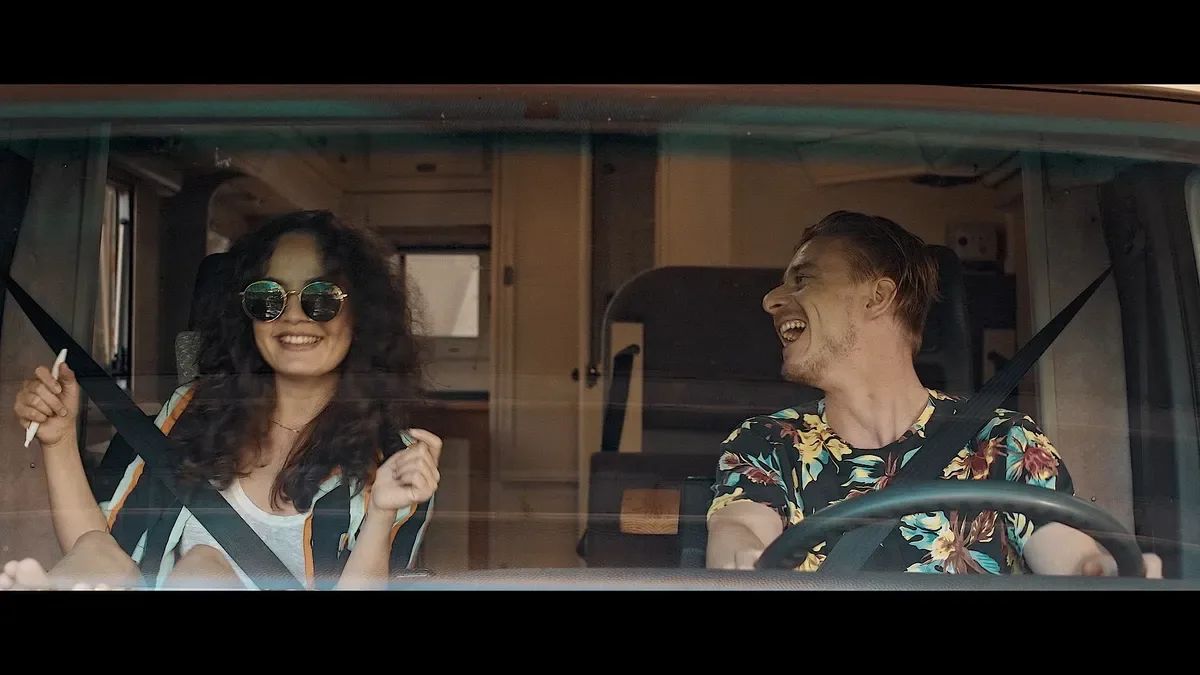 Two young leads laughing in a motorhome cab, framed through the windshield — a woman in round sunglasses on the left, a man in a tropical-print shirt at the wheel