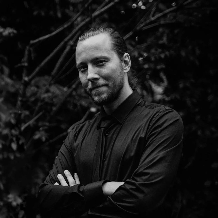 Tamir Gostiša in a black-and-white portrait, arms crossed in a black shirt against a dark woodland backdrop