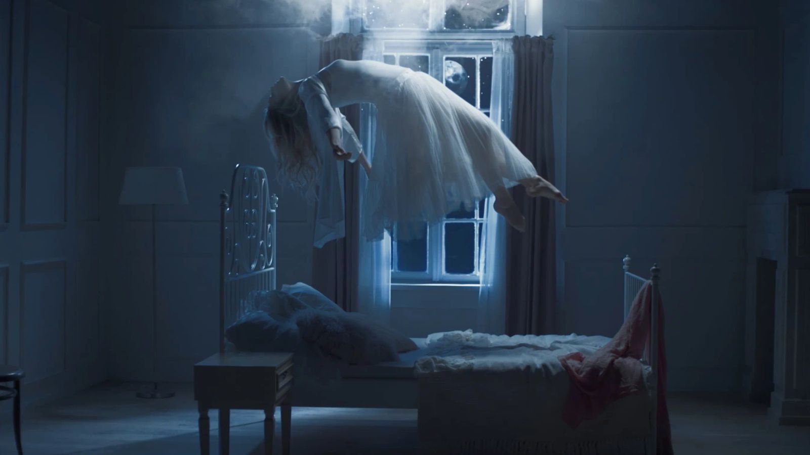 Actress Katarina Cas in a white dress floats above a bed in a moonlit bedroom, drifting toward a glowing window in a dreamlike blue-tinted scene