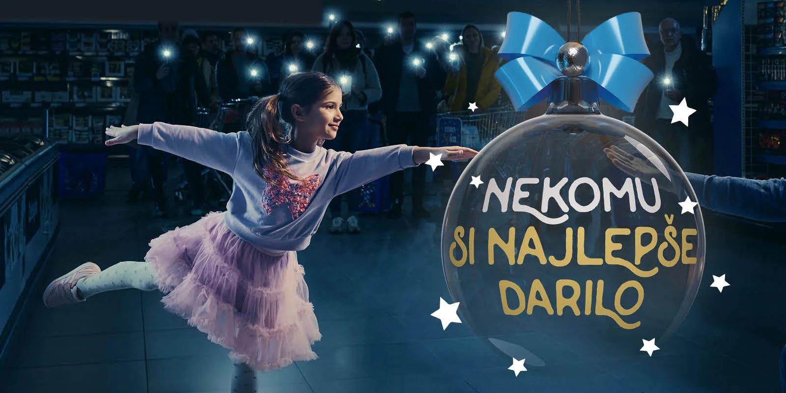 A young girl in a pink tutu runs with arms outstretched toward a bearded man kneeling with open arms inside a darkened supermarket, separated by a giant Christmas bauble carrying the Slovenian campaign line "Nekomu si najlepše darilo"